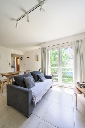 Ein modernes Wohnzimmer mit einem grauen Sofa, einem Esstisch und einem großen Fenster mit Vorhängen.