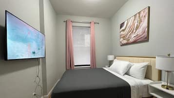 Un dormitorio con cama, televisor montado en la pared, mesita de noche con lámpara y ventana con persianas y cortinas.