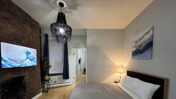 A bedroom with a bed, a TV mounted on a brick wall, a fireplace, and a hallway leading to another room. - Grand Upper West Side Suite, Sleeps 8+, Steps to Central Park (New York)