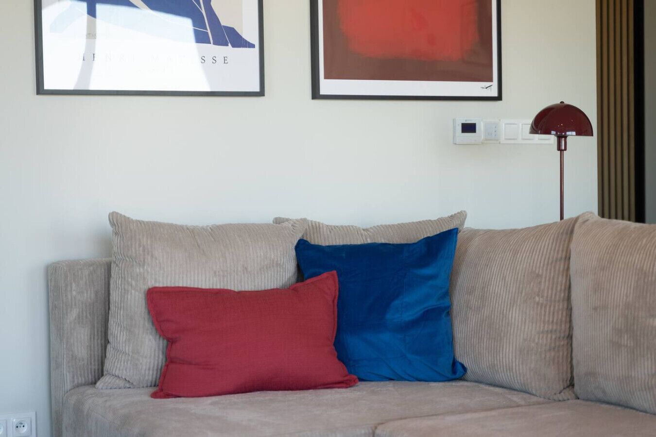 Un sofá color beige con dos cojines, uno rojo y otro azul, y una lámpara de pie roja.