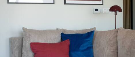 Un sofá color beige con dos cojines, uno rojo y otro azul, y una lámpara de pie roja.