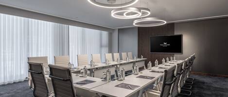 A conference room with a long table, multiple chairs, a flat-screen TV, and circular ceiling lights.