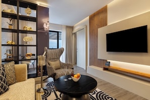 A modern living room with a sofa, armchair, and a round coffee table with fruit and a glass. A flat-screen TV is mounted on the wall, and there's a bookshelf with decorative items. - The Encore Hotel Nakuru (Nakuru)
