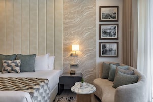 Habitación de hotel con cama, mesita de noche con lámpara, una pequeña mesa redonda con una planta, un sofá y tres cuadros en la pared.
