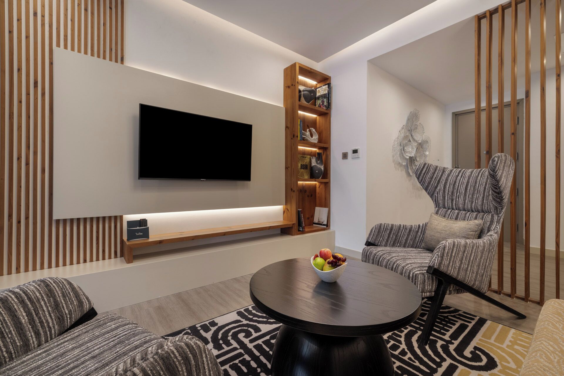 A modern living room with a flat-screen TV, a wooden bookshelf, a round coffee table with fruit, striped sofas, and a patterned rug.