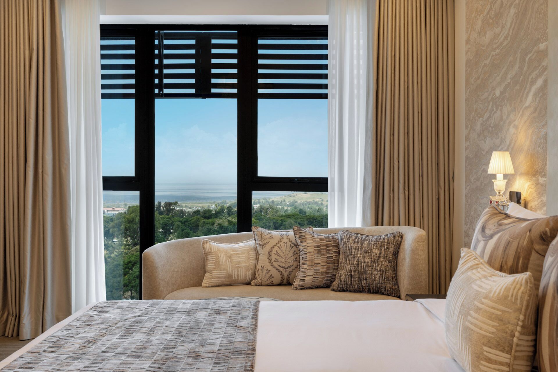 A hotel room with a large bed, a view of a landscape through a window with blinds, and a nightstand with a lamp.