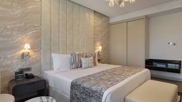 A hotel room with a bed, bedside tables, a desk, a chair, a bench, a chandelier, and a minibar.