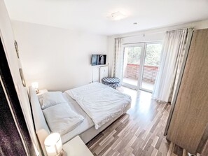 A bedroom with a bed, a TV, a small table, and a balcony with curtains.