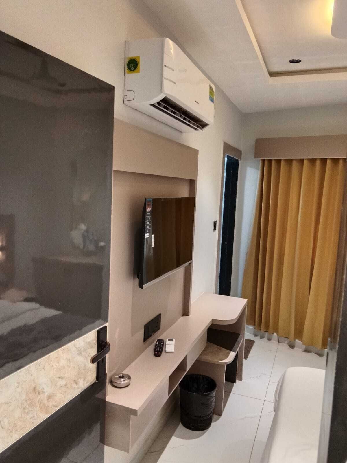 Una habitación de hotel moderna con televisor de pantalla plana, aire acondicionado, un pequeño escritorio, un cubo de basura y un gran ventanal con cortinas.