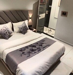 A modern bedroom with a large bed, a bedside table with a lamp, and a glass door leading to a bathroom. - Hotel Bellemount (Ahmedabad)