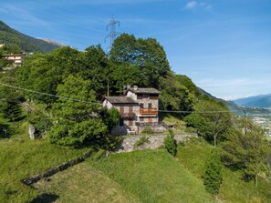 A stone house with a wooden balcony, surrounded by lush greenery and mountains in the background.