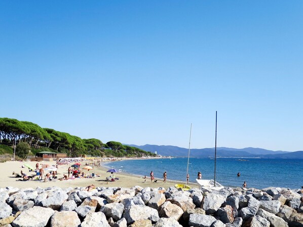 Una playa con primer plano rocoso, gente tomando el sol y barcos en el agua.