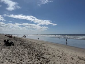 A beach with people walking and sitting on the sand, a clear sky, and the ocean in the background.