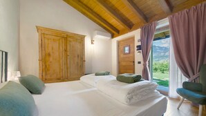 A bedroom with a wooden ceiling, a large bed with white linens, a green chair, and a view of mountains through the window.