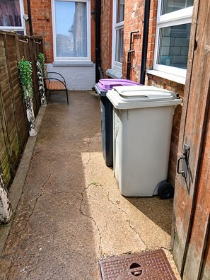 A narrow outdoor pathway leading to a house with a brick exterior, a wooden fence, and a metal trash bin.
