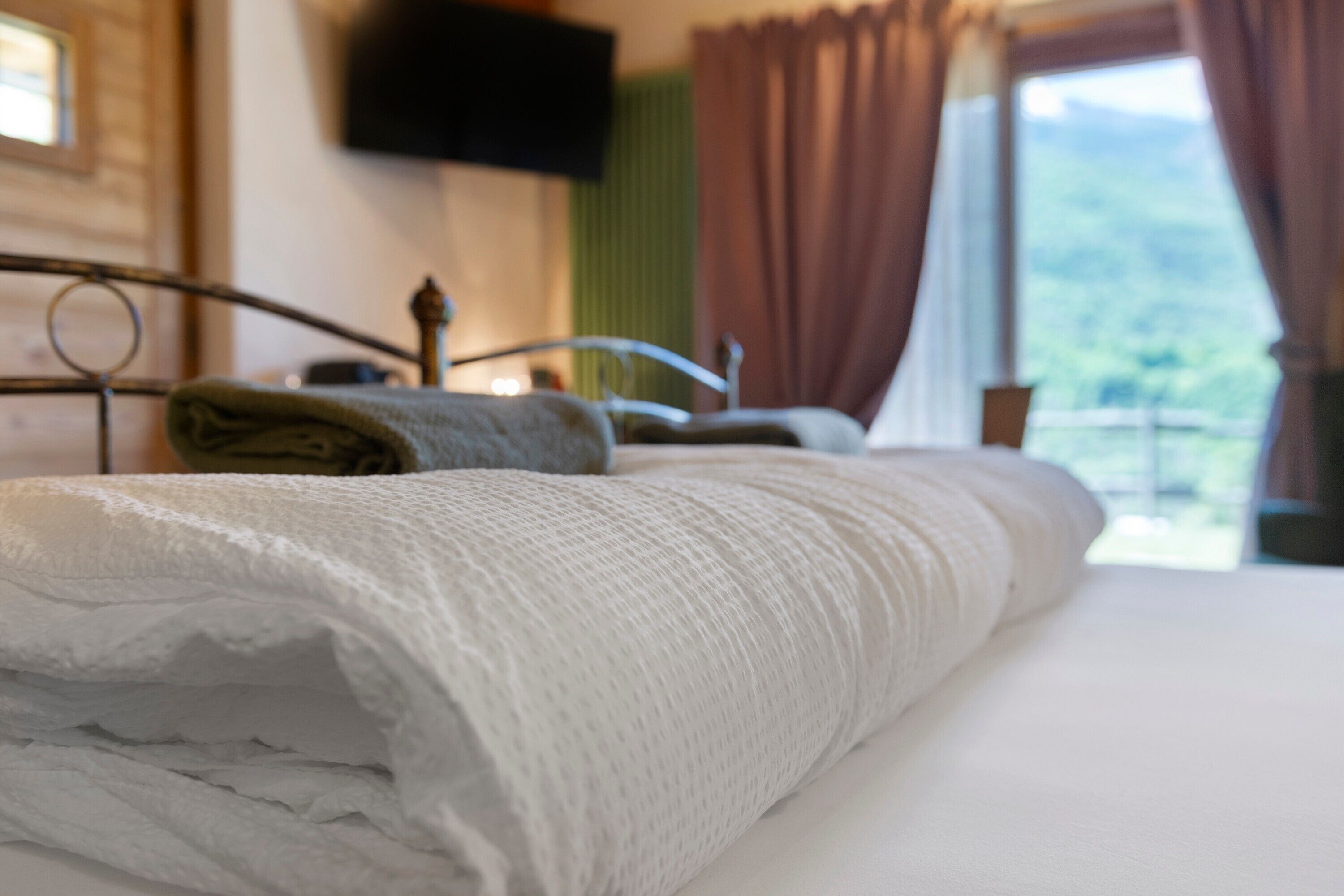 Una cama bien tendida con una manta blanca, estructura de cama de metal, un televisor montado en la pared y una ventana con cortinas.