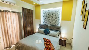Une chambre à coucher comprenant un lit, un ventilateur de plafond, une fenêtre agrémentée d’un élément décoratif au-dessus de la tête de lit, et une table de chevet avec une lampe.