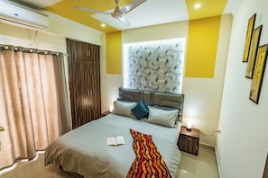 A bedroom with a bed, a ceiling fan, a window with a decorative headboard, and a bedside table with a lamp. - Santa Fe 1BHK CityView by Homeyhuts (Dehradun)