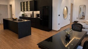 A modern kitchen with black cabinetry, a central island, and a dining area with a glass table. - Terracotta Sunset Suites (Oia)