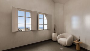A room with two open windows, a round chair, a small wooden table, and a tall vase with sticks. - Terracotta Sunset Suites (Oia)