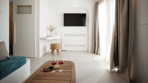 Ett modernt hotellrum med en platt-TV, en soffa, ett soffbord i trä med vinglas och snacks, ett litet sidobord med en vas och ett stort fönster med gardiner.