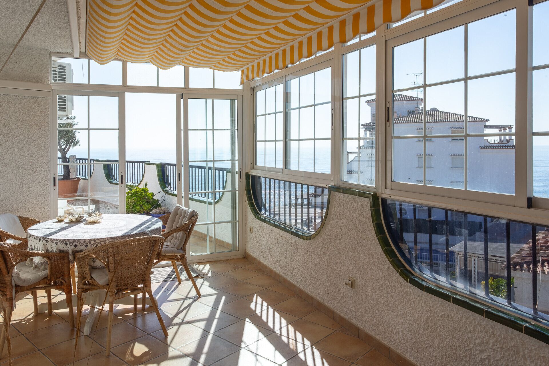 A sunlit room with a table and chairs, large windows with a view of the sea, and a striped awning.