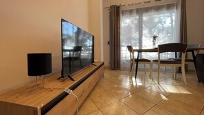 Ein Wohnzimmer mit Fernseher auf einem Holzboden, einem Esstisch mit Stühlen und Blick auf Bäume durch ein Fenster.