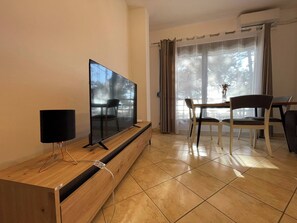 Ein Wohnzimmer mit Fernseher auf einem Holzboden, einem Esstisch mit Stühlen und Blick auf Bäume durch ein Fenster.