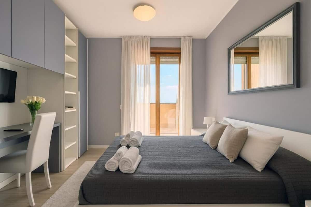 A modern bedroom with a bed, a desk, a TV, and a large window.