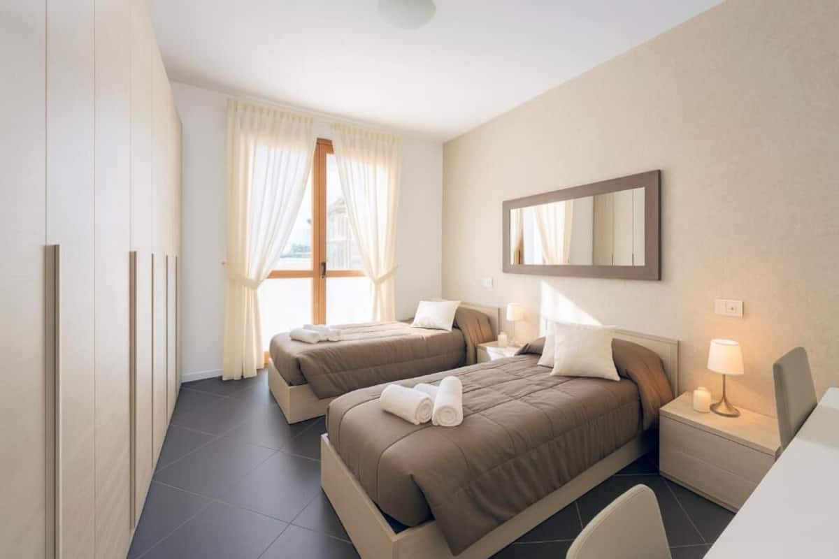 A hotel room with two beds, a large window with curtains, a mirror, and a bedside table with a lamp.