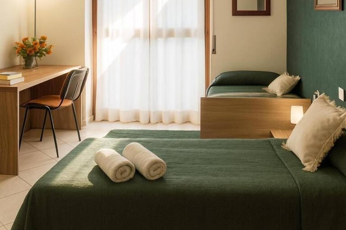 A hotel room with a bed, a desk, a chair, a lamp, and a window with curtains.