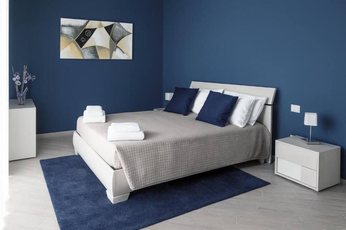 A bedroom with a bed, nightstands, a blue rug, a wall art piece, and a vase with flowers.