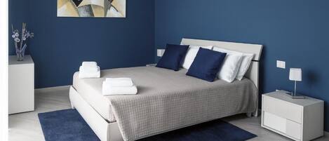 A bedroom with a bed, nightstands, a blue rug, a wall art piece, and a vase with flowers.