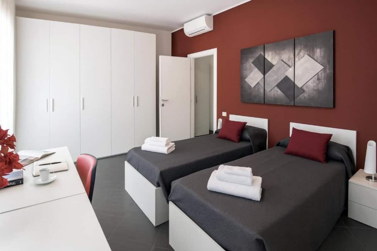 A hotel room with two beds, a desk, a wardrobe, and a painting on the wall.
