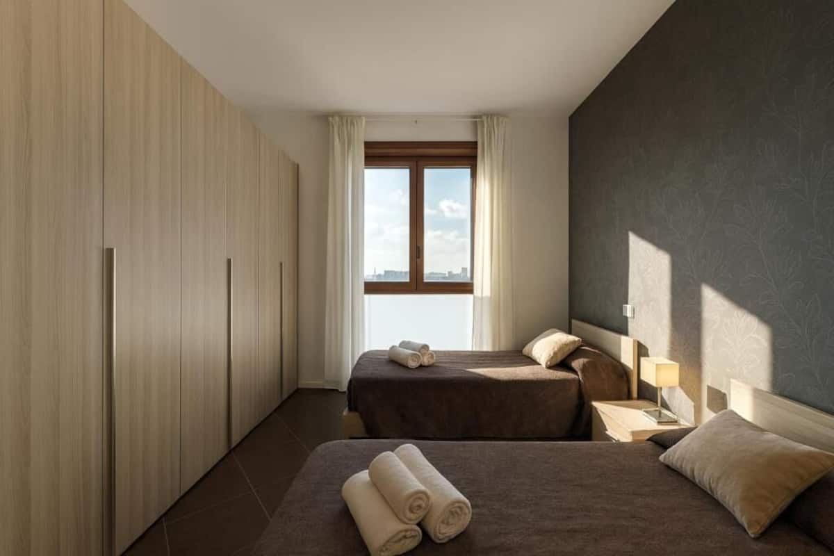 A hotel room with two beds, a large window with curtains, a wardrobe, and a nightstand with a lamp.