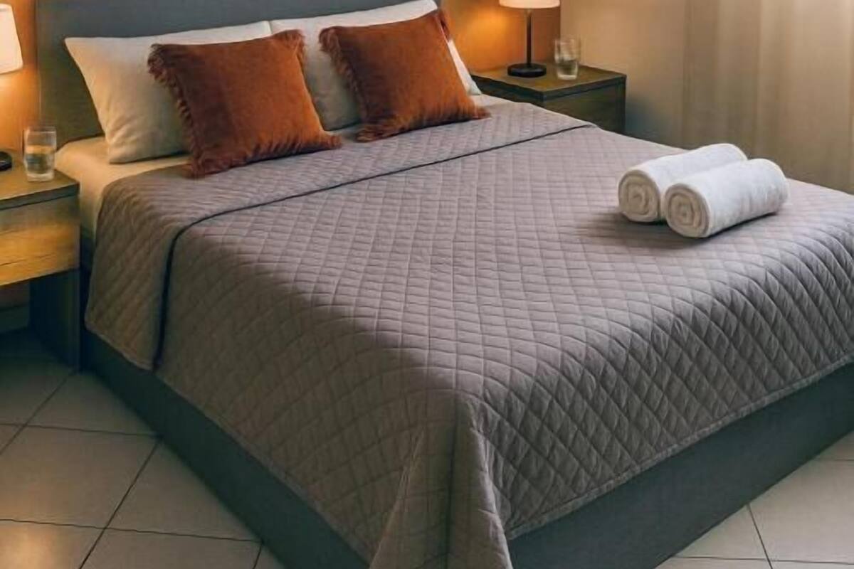 A neatly made bed with a quilted blanket, two pillows, a bedside lamp, a nightstand, and a mirror on the wall.