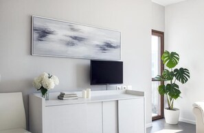 Ein modernes Wohnzimmer mit Fernseher, einem großen abstrakten Gemälde, einem weißen Konsolentisch mit Büchern und Kerzen, einer Topfpflanze und einem Fenster mit Tageslicht.