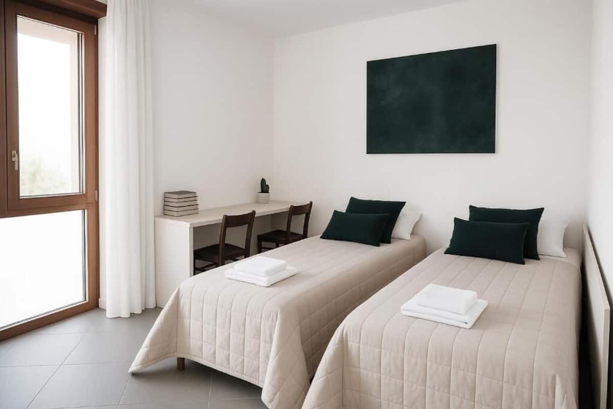 A room with two beds, a desk, a chair, a window with curtains, and a dark-colored artwork on the wall.