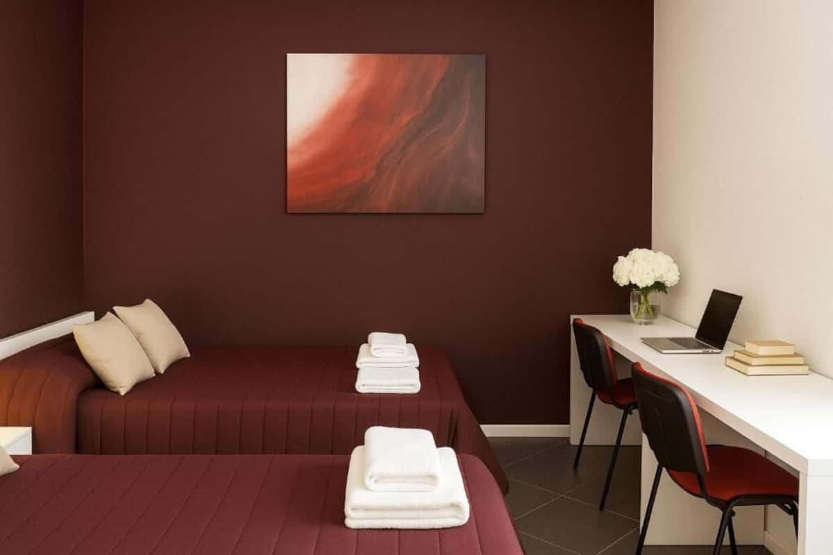 A hotel room with two beds, a desk with a laptop, a vase with flowers, and a painting on the wall.