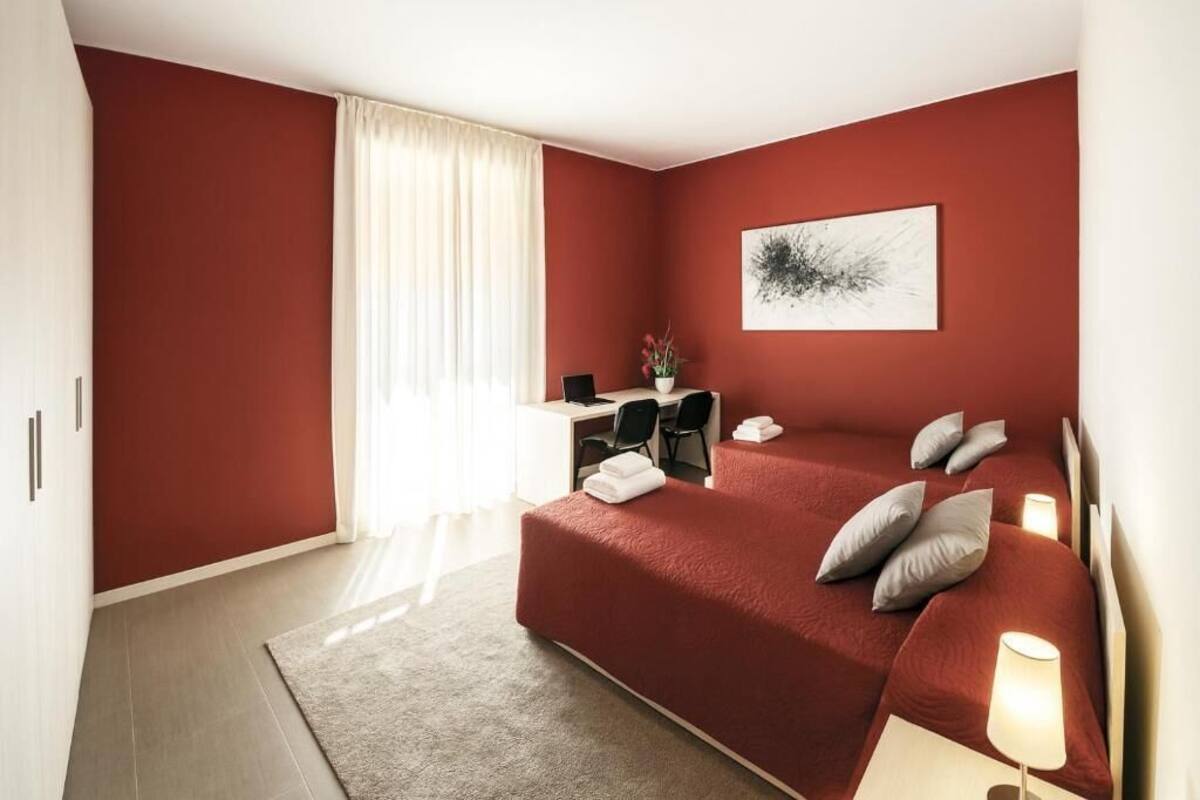A hotel room with a red wall, a bed with pillows, a desk with a chair, and a window with curtains.