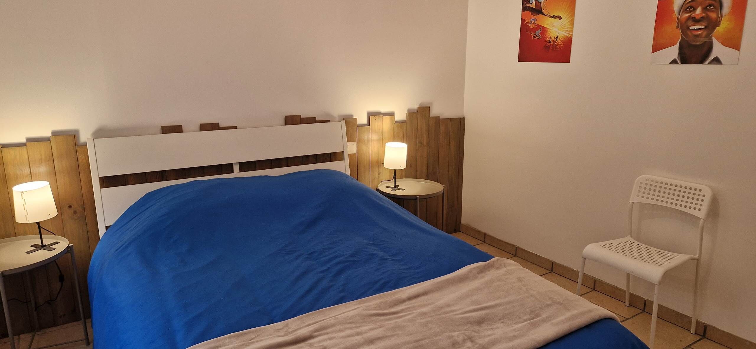 Un lit recouvert d’une couverture bleue, deux lampes de chevet, une chaise blanche et une tête de lit en bois.