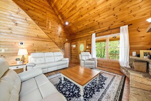 Ein Blockhauskamin mit Holzbalkendecke, einem Sofa, einem Sessel, einem Couchtisch und einem Fenster mit Vorhängen.