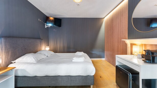 Quarto de hotel moderno com cama, mesa de cabeceira, televisão e uma pequena cozinha.