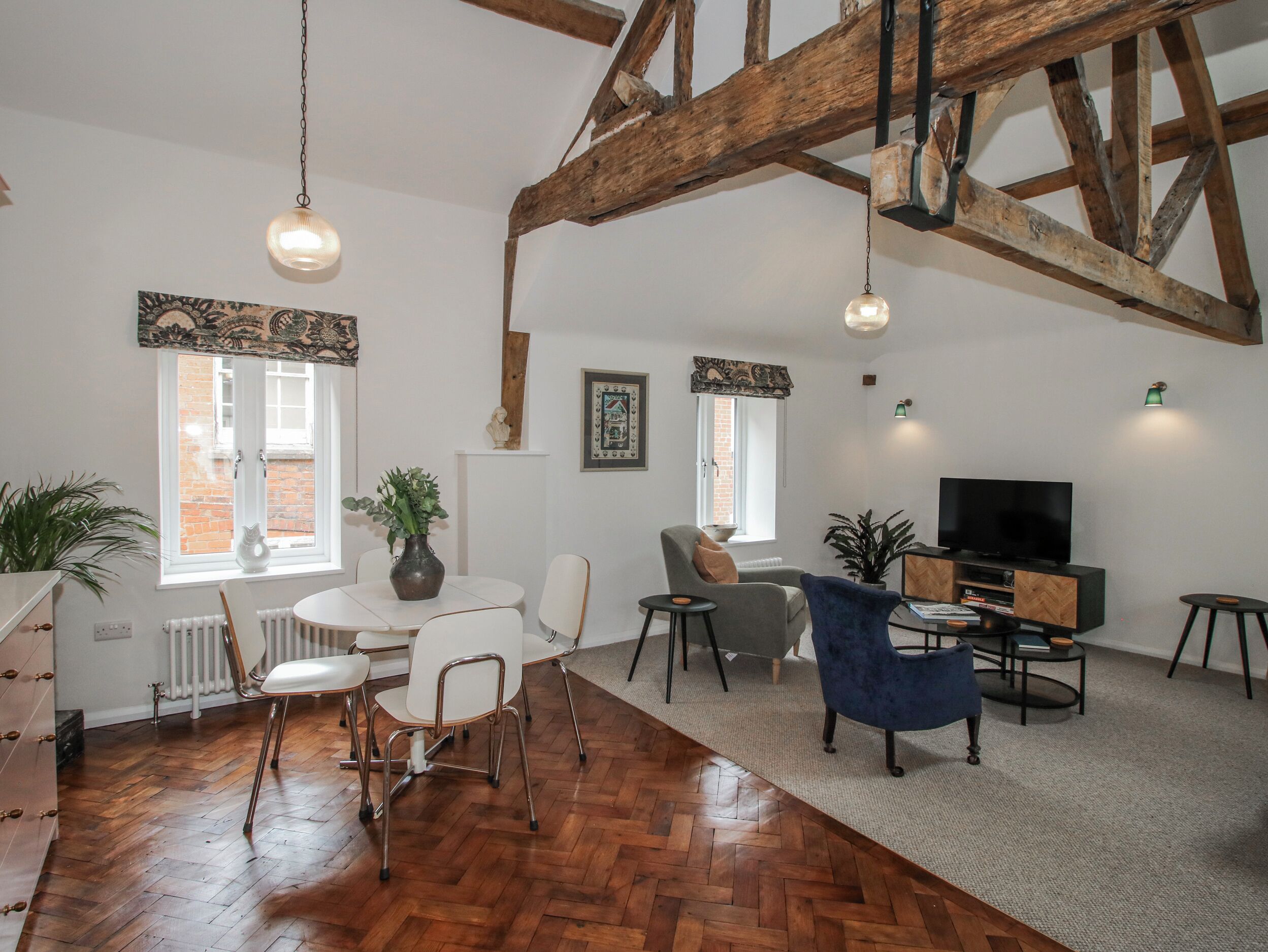Un salón espacioso con suelo de espiga, vigas de madera, chimenea, televisor y comedor con sillas y mesa.