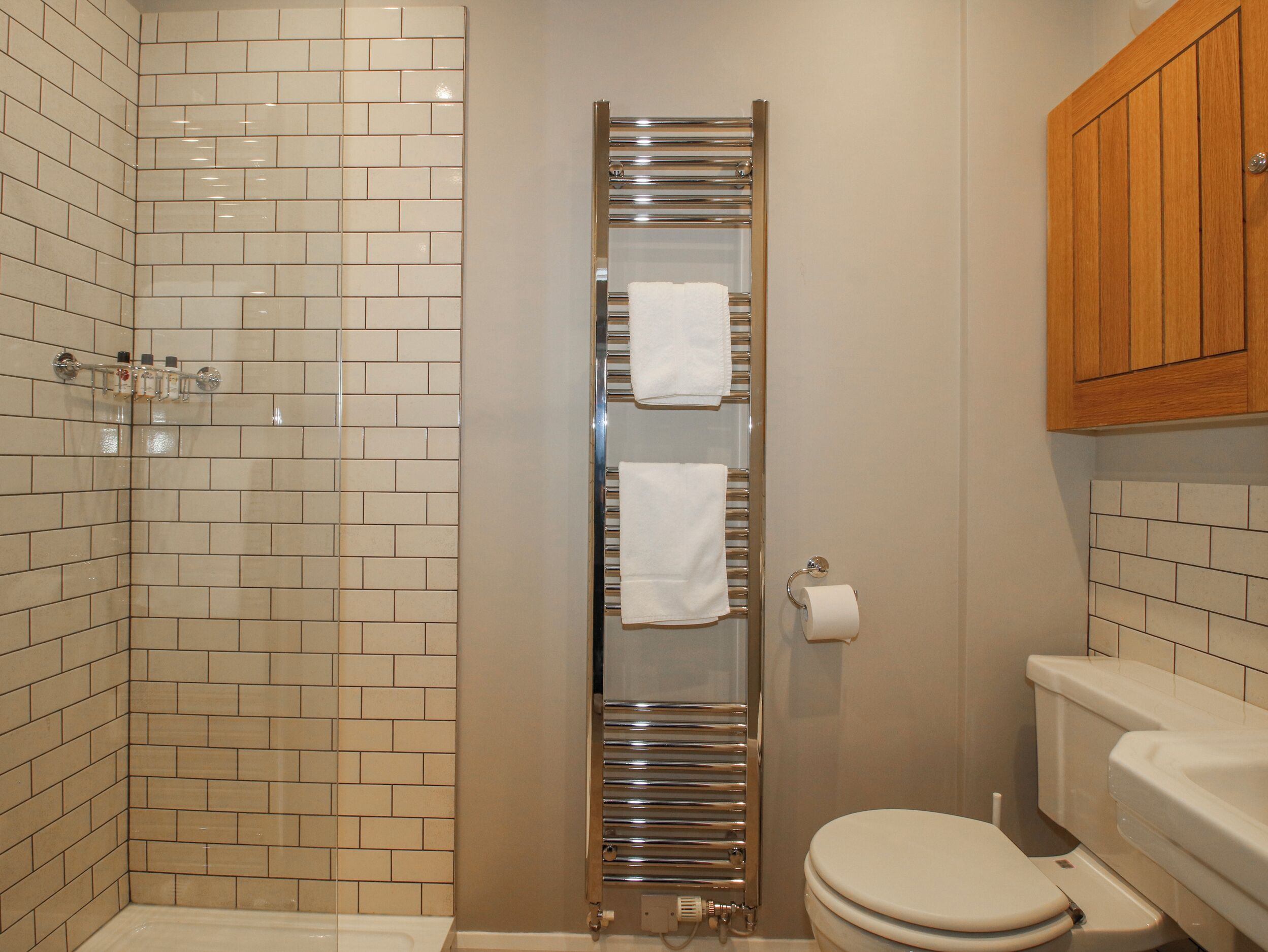 Un baño con pared de ducha azulejada, un toallero vertical, un inodoro y un armario de madera.