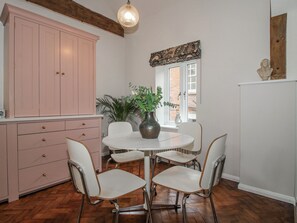 A dining area with a round table, four chairs, a vase with a plant, a pink cabinet, and a window with a view of a brick building. - The Old China Shop (Ludlow)