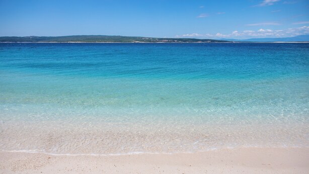 Platja amb aigua cristal·lina, sorra daurada i terra a la distància.