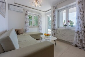 A modern living room with a sofa, a small round table with fruit, and large windows with curtains. - Modern apartment with fantastic balcony and swimming pool (Crikvenica)