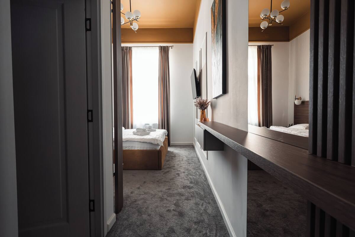 A hotel room with a bed, a bedside table, a mirror, a wall-mounted TV, and a window with curtains.