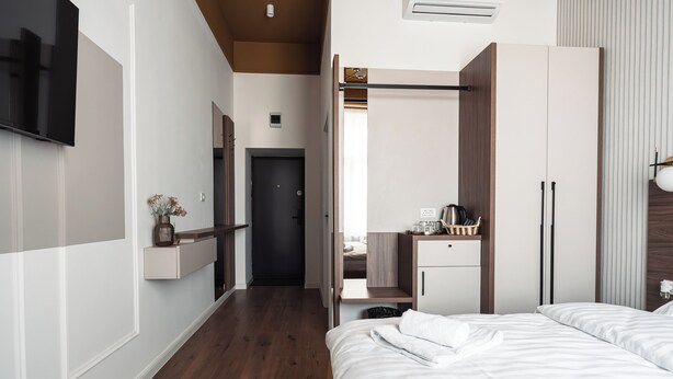 A hotel room with a wooden floor, a bed with white bedding, a TV mounted on the wall, a wardrobe, and a small table with a vase.
